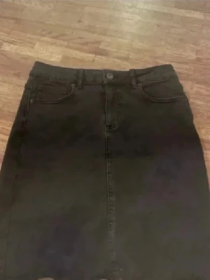 Svart jeanskjol med fickor - Säljer en svart jeanskjol med klassisk femficksdesign och knapp framtill. Kjolen har en rak passform och bälteshällor i midjan. Perfekt att matcha med en t-shirt eller hoodie för en avslappnad look.