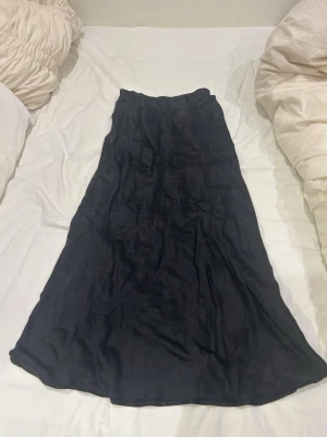 Svart långkjol i satin - Snygg svart långkjol i glansigt satinmaterial. Kjolen har en enkel och stilren design med lätt utsvängd form, perfekt för dig som gillar en klassisk look med modern twist.