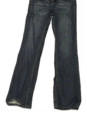 Mörkblå bootcut jeans från Hilfiger - Snöret som syns sitter på insidan då byxorna satt lite löst på mig. Snygga mörkblå bootcut jeans från Tommy Hilfiger med en diskret logga på bakfickan. Jeansen har ljusa slitningar och kontrastsömmar som ger en cool vintagekänsla. Perfekta för dig som gillar en avslappnad men trendig stil. 