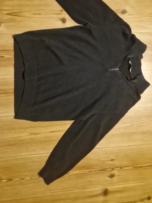 Svart stickad tröja med halv dragkedja - En svart stickad tröja med hög krage och halv dragkedja framtill. Tröjan har långa ärmar och breda ribbstickade muddar vid ärmslut, nederkant och hals. Perfekt för lager-på-lager och enkel att matcha med andra plagg.