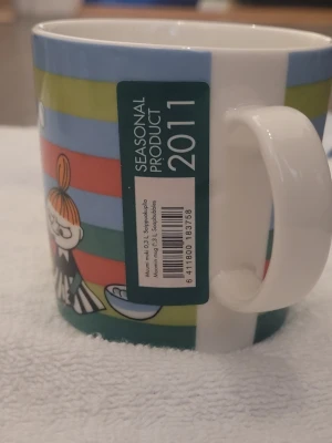 Muminmugg 2011 med Lilla My motiv - Färgglad muminmugg från Arabia med motiv av Lilla My som blåser såpbubblor. Muggen har ränder i blått, grönt, rött och gult och rymmer 0,3 liter. Säsongsprodukt från 2011, tillverkad i porslin.