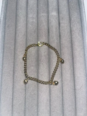 Guldigt armband med hjärtberlocker - Snyggt armband i guldfärgad metall med fyra små hjärtformade berlocker som hänger längs kedjan. Armbandet har en klassisk karbinhake och en chunky kedja som ger en trendig look. Perfekt accessoar för att lyfta din outfit.