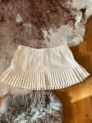 Plisserad vit kortkjol - Superfin vit kortkjol med plisserad nederdel och stilren design. Kjolen har hällor för skärp och är perfekt för att skapa en trendig look. Passar till många olika outfits och är lätt att matcha med både boots och sneakers.