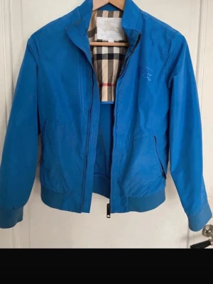 Blå vindjacka från Burberry - Snygg blå vindjacka från Burberry med klassiskt rutigt foder och diskret logga på bröstet. Jackan har dragkedja framtill, två sidofickor med dragkedja. Perfekt för dig som vill ha en jacka till våran.