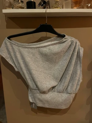 Grå oneshoulder topp  - Snygg ljusgrå oneshoulder topp från Massimo Dutti i mjukt bomullsmaterial. Toppen har bred mudd nedtill och en avslappnad, loose passform som ger en cool siluett. Perfekt att styla med höga jeans eller kjol.