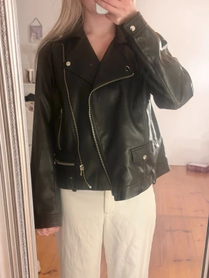 Svart skinnjacka med dragkedjor - Snygg svart skinnjacka i klassisk bikerstil med silvriga dragkedjor och knappar. Jackan har snedställd dragkedja framtill, fickor med dragkedja och tryckknapp samt bred krage. Perfekt för en cool och tidlös look.