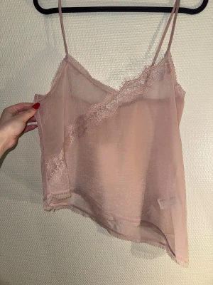 Rosa mesh linnetopp med spets - Säljer en ljusrosa, transparent linnetopp i mesh med tunna axelband och dekorativa spetsdetaljer längs urringningen. Toppen är ärmlös och har en luftig, loose passform som ger en romantisk vibe. Perfekt för lager-på-lager eller som statement-piece.