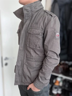 Grå utilityjacka från Superdry - Snygg grå utilityjacka från Superdry med fyra stora fickor framtill, axelklaffar och hög krage. Jackan har patch med brittisk flagga på ärmen och stängs med både dragkedja och knappar. Perfekt för dig som gillar en avslappnad men stilren look.