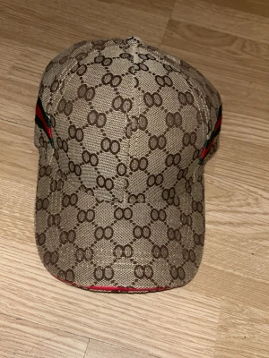 Gucci beige keps med GG-mönster - Säljer en beige keps från Gucci med det klassiska GG-monogrammet över hela kepsen. På sidan finns en grön och röd rand som ger extra lyxig känsla. Kepsen är tillverkad i canvasmaterial och har böjd skärm. Perfekt för dig som vill ha en ikonisk och trendig accessoar. OBS FAKE
