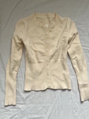 Beige ribbad kofta med knappar - Snygg beige kofta med ribbade detaljer och rund halsringning. Koftan har långa ärmar och stängs med vita knappar framtill. Materialet är mjukt och stretchigt, perfekt för lager på lager. Enkel och stilren design som passar till det mesta.