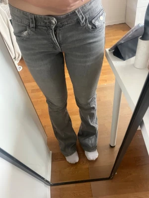 Grå bootcut jeans med detaljer - Säljer ett par grå bootcut jeans med snygga broderade detaljer på bakfickorna och silverfärgade knappar. Jeansen har klassisk femficksmodell och är tillverkade i mjukt denimtyg med lite stretch. Perfekta för dig som gillar en avslappnad men trendig look.