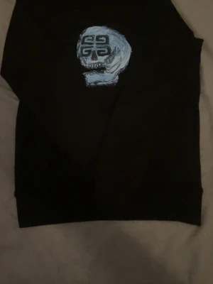 Svart hoodie från Givenchy med dödskalle - Svart hoodie från Givenchy med en stor blå dödskalle och Givenchy-logga på bröstet. På ryggen finns en liten blå Givenchy-text. Hoodien har klassisk passform och är tillverkad i mjukt material, perfekt för en streetwear-look.