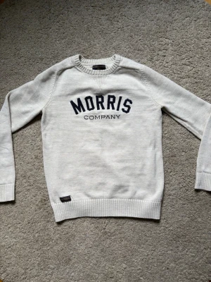 Vit stickad tröja från Morris Gents - Snygg vit stickad tröja från Morris Gents med mörkblå text 'MORRIS COMPANY' på bröstet. Tröjan har rund hals, ribbade muddar och en klassisk passform. Perfekt för dig som gillar stilrena och tidlösa plagg.
