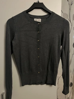 Mörkgrå kofta från Holly & Whyte - Snygg mörkgrå kofta från Holly & Whyte by Lindex med rund halsringning och knappar framtill. Långärmad modell i mjukt material, perfekt att slänga över en topp. Klassisk och enkel design som passar till det mesta.