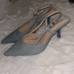 Ljusblå denim spetsiga slingback pumps - Säljer ett par ljusblå pumps med spetsig tå och öppen häl. Skorna har en smal rem med guldfärgat spänne runt ankeln och är klädda i ett tyg som liknar denim. Klackarna är smala och eleganta, perfekta för att lyfta vilken outfit som helst.