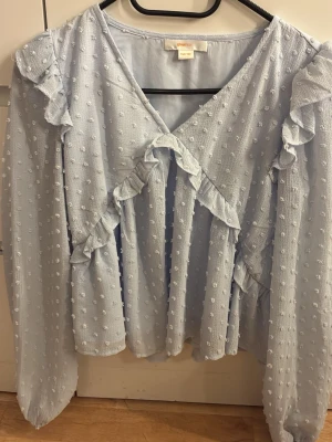 Ljusblå volangblus med prickar - Söt ljusblå blus från Gina Tricot med volangdetaljer och strukturprickar i tyget. Blusen har v-ringning och långa ärmar samt en luftig passform. Perfekt för dig som gillar romantisk och trendig stil.