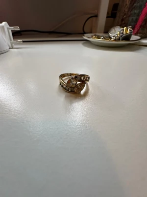 Guldfärgad ring med stenar - Snygg guldfärgad ring med en större klar sten i mitten och mindre stenar längs bandet. Ringen har en unik, böjd design som ger den en modern och cool känsla. Perfekt accessoar för att lyfta din stil.