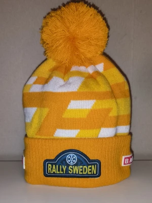 Ovanlig gul Rally Sweden mössa med tofs - Säljer en gul och vit stickad mössa med stor tofs på toppen. Mössan har ett grafiskt mönster, uppvikt kant och broderade märken från Rally Sweden och Bauhaus. Perfekt för dig som gillar motorsport och vill sticka ut i vinterkylan. Mössan säljs inte på någon hemsida eller affär!