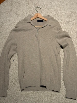 Beige stickad tröja med dragkedja Riley - Snygg beige stickad tröja från Riley med halv dragkedja och krage. Tröjan har lång ärm och är tillverkad i ett grovstickat material som ger en avslappnad men stilren look. Perfekt för lager-på-lager och enkel att matcha med jeans eller chinos.