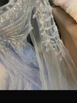 Ljusblå långklänning med pärlor - Magisk ljusblå långklänning med transparenta ärmar och vackra pärl- och paljettbroderier över hela livet. Klänningen har ett svepande tyg i flera lager och detaljer som ger en riktigt lyxig känsla.