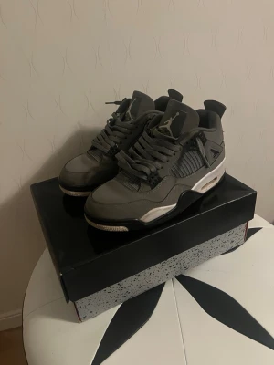 Gråa Air Jordan 4 Retro sneakers - Snygga Air Jordan 4 Retro sneakers i grått med svarta och vita detaljer. Skorna har meshpaneler på sidorna, klassisk snörning och synlig Air-enhet i sulan. Tillverkade i en mix av läder och textil för en clean och sportig look.
