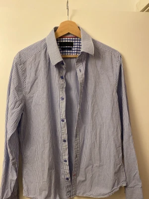 Blårutig skjorta från Riley Slim Fit - Snygg blårutig skjorta från Riley i slim fit-modell. Skjortan har klassisk krage, långa ärmar och är tillverkad i mjuk bomull. Perfekt för dig som gillar en stilren och fräsch look med lite smalare passform. Inte använd. Mycket bra skick