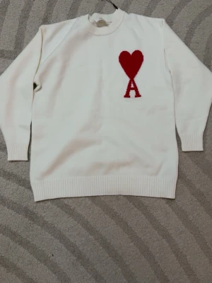 Ami paris tröja - Helt ny vit stickad tröja från Ami med ett rött hjärta och bokstaven A broderat på bröstet. Tröjan har rund halsringning, ribbade muddar och är gjord i mjukt material. Perfekt för en stilren och trendig look.