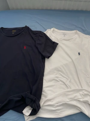 2-pack t-shirts från Polo Ralph Lauren - Säljer två klassiska t-shirts från Polo Ralph Lauren. En marinblå med röd broderad logga och en vit med marinblå broderad logga. Båda är kortärmade och i mjuk bomull, perfekt basic till garderoben.