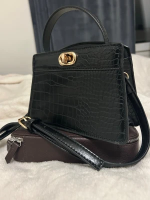Svart croco handväska med gulddetaljer - Snygg svart handväska i croco-mönstrat konstläder med guldfärgat vridlås och matchande detaljer. Väskan har både handtag och justerbar axelrem, perfekt för att bära på olika sätt. Stilren och trendig accessoar som lyfter din outfit.