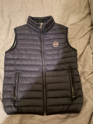 Svart dunväst från Colmar - Snygg mörkblå dunväst från Colmar med quiltad design och hög krage. Västen har två dragkedjeförsedda fickor framtill och Colmar-logga på bröstet. Perfekt att slänga över en hoodie eller tröja för extra värme. Materialet är lätt och smidigt.