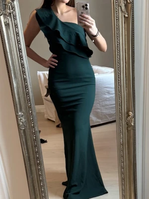 Mörkgrön oneshoulder långklänning - Elegant mörkgrön långklänning med oneshoulder-design och dubbla volangdetaljer över axeln. Klänningen har en figurnära passform och är perfekt för speciella tillfällen. Materialet känns mjukt och följsamt.
