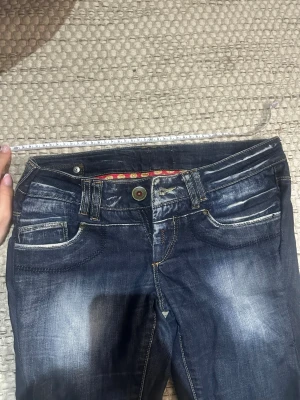 Mörkblå bootcut jeans med slitningar - Säljer ett par mörkblå bootcut jeans med slitna detaljer och råa kanter nertill. Jeansen har klassisk femficksdesign, kontrastsömmar och en cool tvätt med ljusare partier. Perfekta för dig som gillar en avslappnad och trendig stil. midjemått:39cm innerben:83cm    
