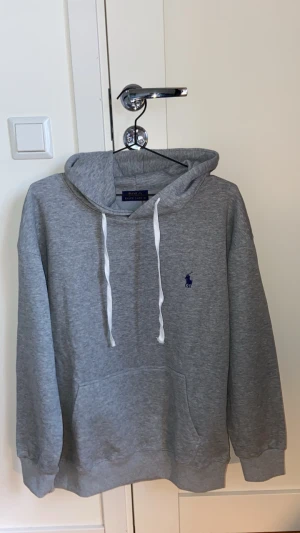 Grå hoodie från Polo Ralph Lauren - Klassisk grå hoodie från Polo Ralph Lauren med blå broderad logga på bröstet. Hoodien har vit dragsko, känguruficka och ribbade muddar. Perfekt för en avslappnad och stilren look. Tröjan är helt oanvänd! Hör av er kring frågor😁