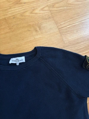 Mörkblå tröja från Stone Island - Snygg mörkblå tröja från Stone Island med rund halsringning och ribbade muddar. Tröjan har lång ärm och den klassiska Stone Island-loggan på vänster ärm. Materialet är ribbstickat och ger en stilren look.