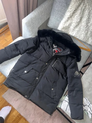 Svart dunjacka Moose Knuckles XL - Snygg svart dunjacka från Moose Knuckles med stor huva och svart fuskpäls. Jackan har flera fickor med tryckknappar, dragkedja och logotyp på ärmen. Perfekt för kalla vinterdagar och riktigt varm.