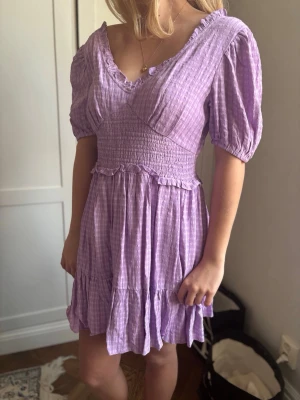 Lila rutig kortklänning med puffärm - Superfin lila klänning med rutigt mönster och v-ringad hals. Klänningen har smock i midjan, volangdetaljer och puffiga korta ärmar. Perfekt för dig som gillar en feminin och trendig stil. Materialet känns lätt och luftigt.