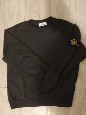Svart Sweatshirt Stone Island Storlek L - Svart sweatshirt från Stone Island med klassisk rund hals och ikonisk logopatch på vänster ärm. Tröjan har ribbade muddar vid ärmslut och nederkant. Perfekt för en avslappnad och stilren look.