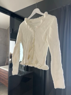 Vit fluffig kofta med volangkant - Supermjuk och fluffig vit kofta med volangdetaljer längs knäppningen och nederkanten. Långärmad och figurnära modell som ger en söt och trendig look. 