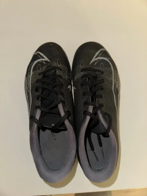 Nike svart/gråa fotbollsskor - Snygga fotbollsskor från Nike i svart med grå detaljer och vit Nike-logga på sidan. Skorna har snörning och en låg profil, perfekt för snabba rörelser på planen. Ovandelen är i syntetmaterial för bra bollkontroll.
