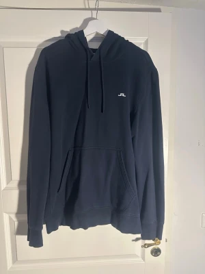 Mörkblå hoodie från J.Lindeberg - Snygg mörkblå hoodie från J.Lindeberg med klassisk känguruficka och liten vit logga på bröstet. Hoodien har dragsko i huvan och ribbade muddar vid ärmar och nederkant. Perfekt för en chill och stilren look. Den är i mindre i storleken så mer en L/XL storlek