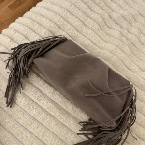 Grå mocka kuvertväska med fransar - Säljer en snygg grå kuvertväska i mjuk mocka med långa fransar på sidorna. Väskan har en enkel och stilren design med knytband framtill. Perfekt accessoar för att lyfta din outfit.