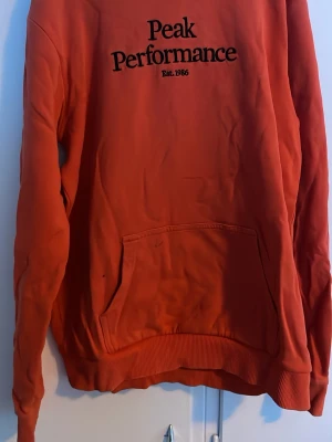 Röd hoodie från Peak Performance - Snygg röd hoodie från Peak Performance med svart logga och text 'Est. 1986' på bröstet. Klassisk känguruficka framtill och ribbade muddar vid ärmar och nederkant. Perfekt för en avslappnad och sportig stil.