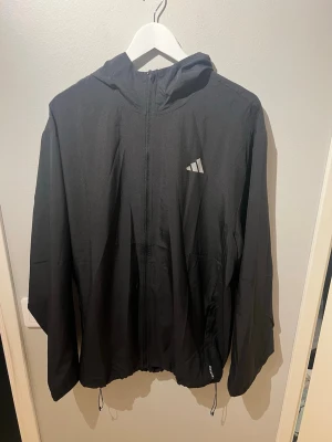 Helt oanvänd svart löparjacka/vindjacka från Adidas - Aldrig använd med prislappen kvar. Säljes på grund av fel storlek.      Snygg svart löparjacka/vindjacka från Adidas med huva och hel dragkedja framtill. Jackan har Adidas-logga på bröstet och ryggen. Tillverkad i lätt och funktionellt material, perfekt för löpning och träning.