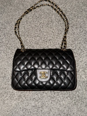 Svart Chanel axelväska i läder - Säljer en klassisk svart axelväska från Chanel i quiltat läder med guldfärgad kedja och ikoniskt CC-spänne framtill. Väskan har tidlös design och elegant form, perfekt för dig som vill ha en lyxig och stilren accessoar.