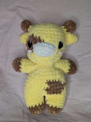Gul och brun virkad giraff - Söt handgjord giraff i mjukt, virkat garn. Färgerna är ljusgul med bruna detaljer på öron, ben, svans och fläckar. Ansiktet har vita och svarta detaljer samt små rosa kinder. Giraffen är 19 cm lång och 14 cm bred med armarna. Perfekt som gosedjur eller dekoration.