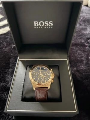 Hugo Boss guld herrklocka brun läder - Stilren herrklocka från Hugo Boss med guldfärgat boett, svart urtavla och brunt läderarmband. Klockan har flera små urtavlor och tydliga markeringar. Perfekt accessoar för dig som gillar klassisk och lyxig stil.