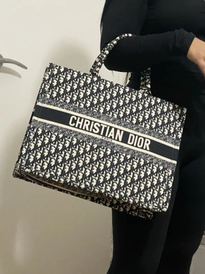 Tote shoppingväska - Snygg och rymlig Book Tote shoppingväska svart och vit canvas med ikoniskt mönster och logga framtill. Väskan har två handtag och en boxig, rektangulär form som rymmer allt du behöver till skolan eller stan.