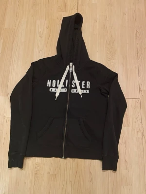 Svart Hollister hoodie med dragkedja - Svart hoodie från Hollister med vit logga och texten 'California' på bröstet. Tröjan har dragkedja framtill, två fickor och vita snören i huvan. Perfekt för en avslappnad och cool stil.