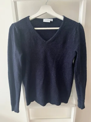Mörkblå stickad tröja från Vila - Mysig mörkblå stickad tröja från Vila med v-ringning och långa ärmar. Tröjan har ribbstickade muddar vid ärmslut och nederkant, vilket ger en snygg och enkel look. Perfekt att matcha med jeans eller kjol för en avslappnad stil. Köparen står för eventuell frakt.