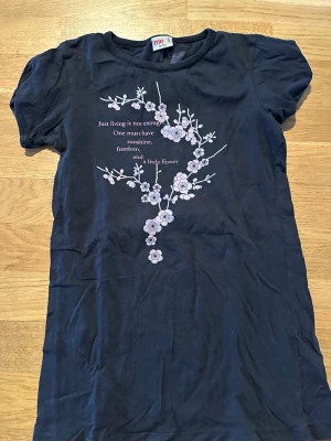 Svart blommig t-shirt med tryck Barn! - Svart t-shirt från Me Too med korta ärmar och ett blommigt tryck framtill samt text. På ryggen finns två små fjärilar i rosa och vitt. T-shirten har en klassisk passform och är gjord i mjuk bomull, perfekt för en avslappnad stil.